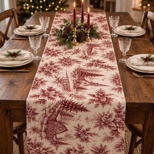 May include: A cream-colored table runner featuring a repeating deep red toile pattern of winter scenes, including houses, church steeples, and pine trees. The fabric is laid across a wooden dining table set with white napkins and clear glassware.