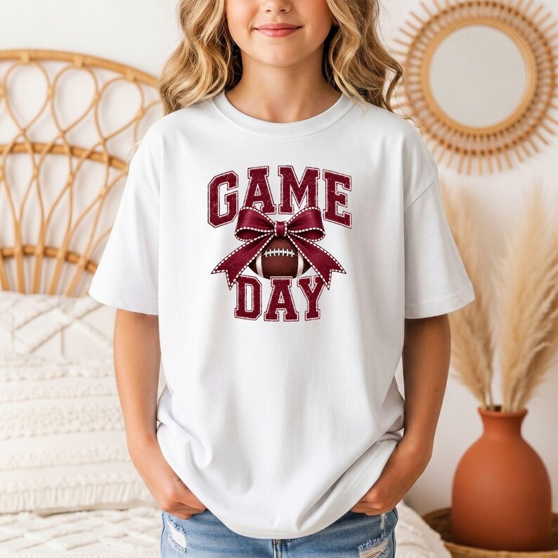 Youth white Comfort Colors Game Day football shirt with a maroon bow and football graphic.