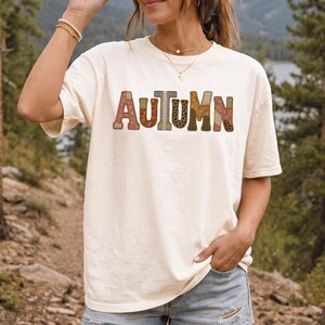 Puede incluir: Una camiseta de manga corta de color crema con la palabra AUTUMN impresa en el pecho. Cada letra está rellena con un patrón único, incluyendo texturas florales, de cuadros, de leopardo y tonos tierra sólidos en marrón y naranja.