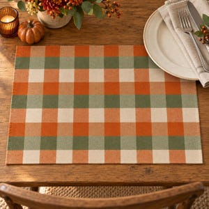 May include: A rectangular placemat featuring a checkered pattern in alternating squares of burnt orange, sage green, and cream. The fabric has a visible woven texture, creating a grid design that covers the entire surface of the rectangular mat.
