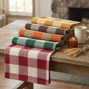 May include: A stack of five rectangular fabric table runners featuring a large buffalo check pattern in red, green, orange, brown, and yellow. Each runner is folded neatly, with the red and white one draped over the edge of a wooden table.