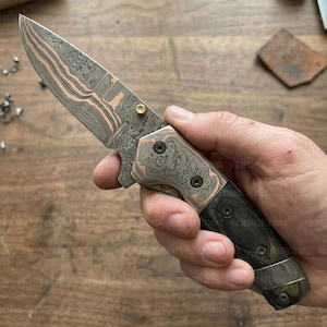 May include: A folding knife with a patterned blade and copper-colored accents. The handle is a dark wood with a copper-colored bolster. The blade has a unique, layered design. The knife is held in a hand.