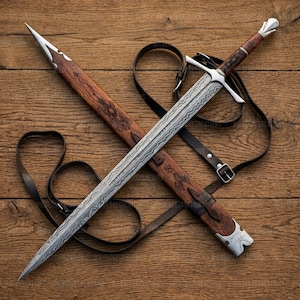 May include: An overhead view of a sword and scabbard. The sword features a silver blade with a patterned design and a brown handle. The scabbard is brown with decorative carvings and silver accents. A black leather strap is attached.