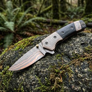 K&ouml;nnte beinhalten: Ein Klappmesser mit gemusterter Klinge und Griff, mit einem Design aus dunklem Holz und kupferfarbenem Metall. Die Klinge hat ein einzigartiges, geschichtetes Muster. Das Messer liegt auf einem moosbedeckten Felsen, mit einem Waldboden.