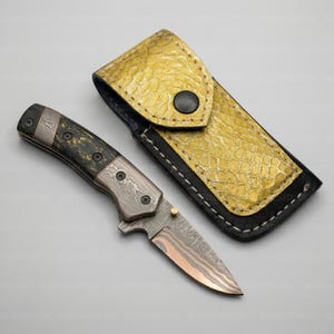K&ouml;nnte beinhalten: Ein Klappmesser mit gemusterter Klinge und dunklem Griff, neben einer goldfarbenen Lederscheide. Die Messerklinge hat einen kupferfarbenen Farbton. Die Scheide ist strukturiert und hat einen schwarzen Knopfverschluss. Das Messer ist etwa 18 cm lang.