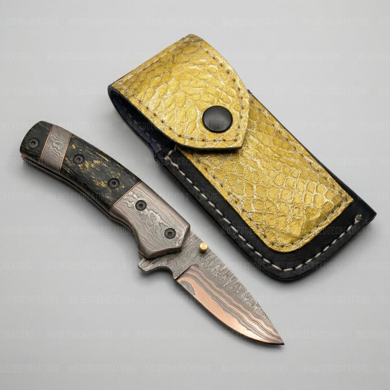 K&ouml;nnte beinhalten: Ein Klappmesser mit gemusterter Klinge und dunklem Griff, neben einer goldfarbenen Lederscheide. Die Messerklinge hat einen kupferfarbenen Farbton. Die Scheide ist strukturiert und hat einen schwarzen Knopfverschluss. Das Messer ist etwa 18 cm lang.
