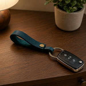 May include: A teal leather keyring with a gold rivet is attached to a car key fob. The key fob is black with silver trim and several buttons. The keyring rests on a dark brown wooden surface.