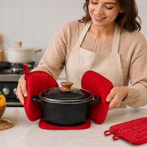 May include: A black cooking pot with a wooden handle is held with red oven mitts and a matching pot holder. A bowl of fresh vegetables and a loaf of bread are on the counter. Kitchenware.