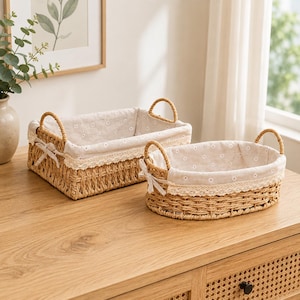 May include: Two woven seagrass baskets with light beige fabric liners featuring a small white daisy pattern. Each basket has two side handles, a decorative lace trim along the top edge, and a small fabric bow tied on one side of the basket rim.