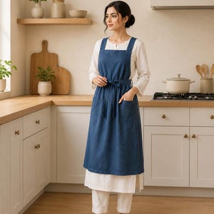 May include: A denim blue apron with a square neckline and a tie waist. The apron has two front pockets and is worn over a white long-sleeved shirt and white pants. The person is standing in a kitchen with wooden countertops and white cabinets.