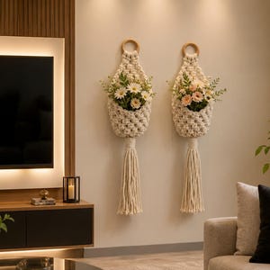 May include: Two off-white macrame wall hangings featuring a woven diamond pattern, a circular wooden ring at the top, and a long tassel at the bottom. Each hanging holds a bouquet of artificial flowers with white, pink, and green foliage.