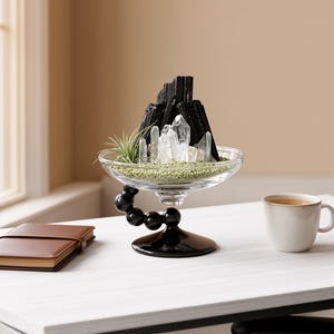 Puede incluir: Un cuenco de cristal transparente con una base de pedestal negra que presenta una serie de esferas apiladas. Dentro del cuenco, una colecci&oacute;n de cristales de turmalina negra y cuarzo transparente se dispone con una peque&ntilde;a planta de aire.