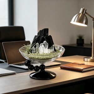 Puede incluir: Un cuenco de cristal transparente sobre una base de pedestal negra con detalles esf&eacute;ricos. En su interior contiene fragmentos de turmalina negra, cristales de cuarzo transparente y una peque&ntilde;a planta de aire sobre grava verde fina.