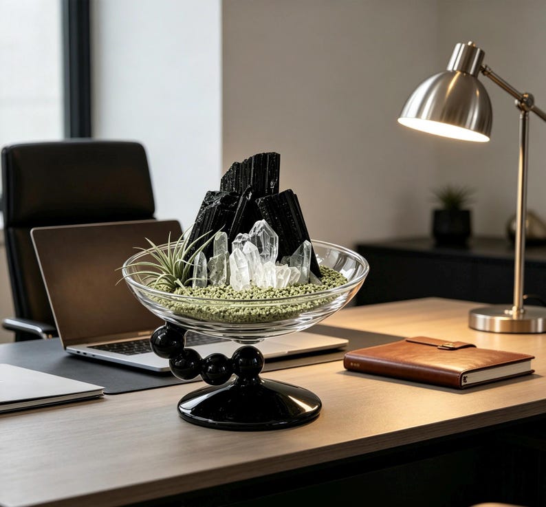 Puede incluir: Un cuenco de cristal transparente sobre una base de pedestal negra con detalles esf&eacute;ricos. En su interior contiene fragmentos de turmalina negra, cristales de cuarzo transparente y una peque&ntilde;a planta de aire sobre grava verde fina.