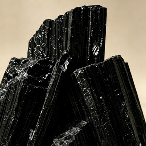 Puede incluir: Un grupo de cristales de turmalina negra en bruto con estr&iacute;as verticales profundas y una superficie brillante y reflectante. La formaci&oacute;n mineral muestra bordes afilados y dentados, junto con un color oscuro y opaco muy caracter&iacute;stico.