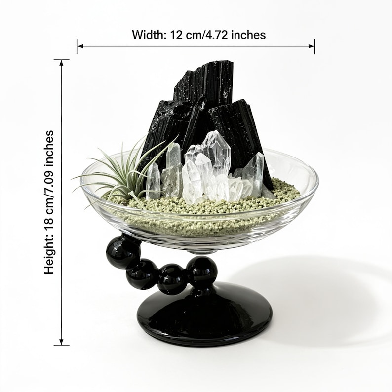 Puede incluir: Un cuenco de cristal transparente sobre una base de pedestal negra con un brazo curvo de esferas negras, que contiene turmalina negra, cristales de cuarzo transparentes y una peque&ntilde;a planta de aire sobre arena verde. Mide 12 cm de ancho y 18 cm de alto.
