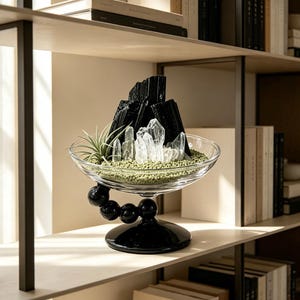 Puede incluir: Un cuenco de cristal transparente con una base de pedestal negra formada por esferas. Dentro del cuenco hay cristales de turmalina negra, puntas de cuarzo transparente, una peque&ntilde;a planta de aire y una capa de grava verde claro decorativa.