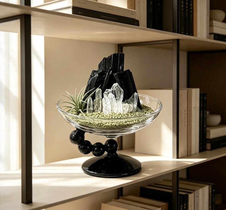 Puede incluir: Un cuenco de cristal transparente con una base de pedestal negra formada por esferas. Dentro del cuenco hay cristales de turmalina negra, puntas de cuarzo transparente, una peque&ntilde;a planta de aire y una capa de grava verde claro decorativa.