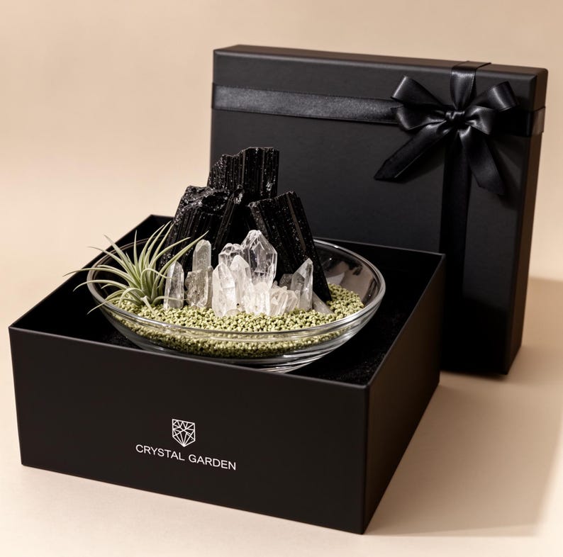 Puede incluir: Un cuenco de cristal transparente con turmalina negra, cristales de cuarzo transparente y una peque&ntilde;a planta de aire sobre grava verde. El cuenco est&aacute; dentro de una caja de regalo negra con un lazo y el texto CRYSTAL GARDEN impreso.