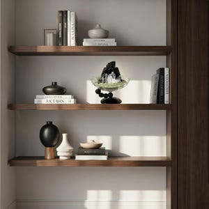 Puede incluir: Tres estantes flotantes de madera oscura que contienen varios objetos decorativos, incluidos libros con lomos blancos y negros, un jarr&oacute;n de cer&aacute;mica gris, un jarr&oacute;n esf&eacute;rico negro, un jarr&oacute;n geom&eacute;trico blanco y un cuenco de cristal con cristales negros.