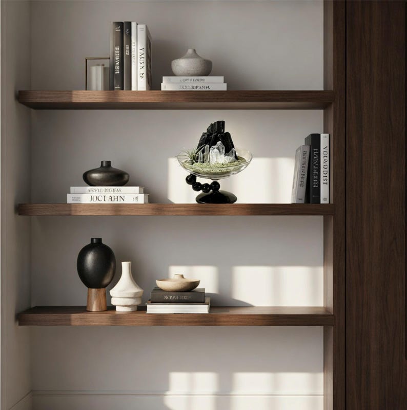 Puede incluir: Tres estantes flotantes de madera oscura que contienen varios objetos decorativos, incluidos libros con lomos blancos y negros, un jarr&oacute;n de cer&aacute;mica gris, un jarr&oacute;n esf&eacute;rico negro, un jarr&oacute;n geom&eacute;trico blanco y un cuenco de cristal con cristales negros.