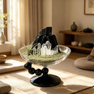 Puede incluir: Un cuenco de cristal transparente con tallo y base esf&eacute;ricos negros, relleno de arena verde, varias puntas de cuarzo transparente, fragmentos de turmalina negra y una peque&ntilde;a planta a&eacute;rea. El cuenco se apoya en un brazo curvo con cuatro esferas.