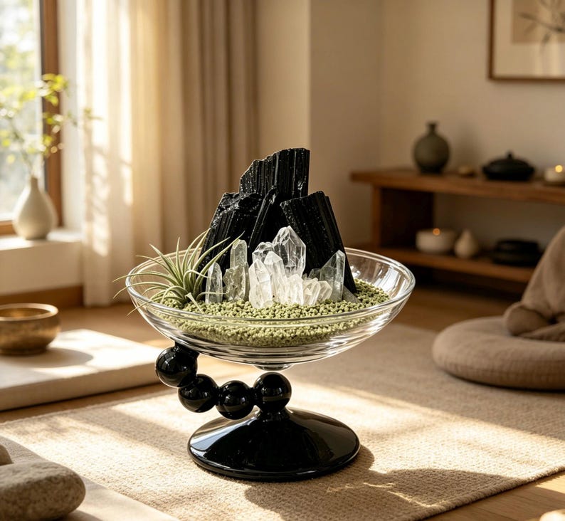 Puede incluir: Un cuenco de cristal transparente con tallo y base esf&eacute;ricos negros, relleno de arena verde, varias puntas de cuarzo transparente, fragmentos de turmalina negra y una peque&ntilde;a planta a&eacute;rea. El cuenco se apoya en un brazo curvo con cuatro esferas.