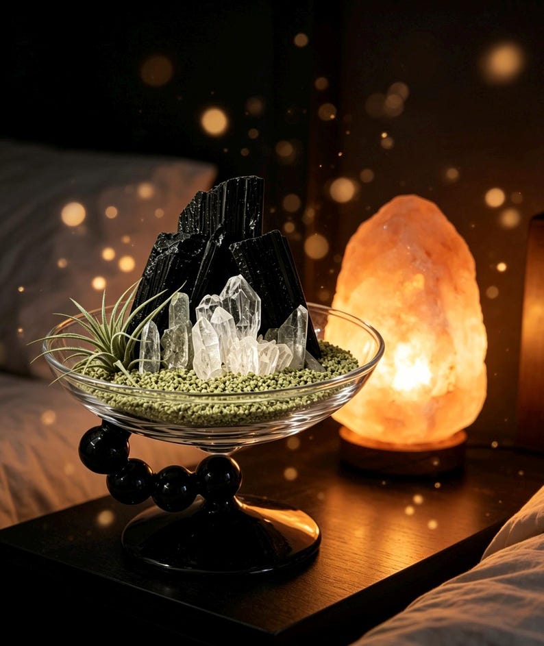 Puede incluir: Un cuenco de cristal transparente con base esf&eacute;rica negra que contiene arena verde, cristales de cuarzo transparente, fragmentos de turmalina negra y una peque&ntilde;a planta de aire. Junto al cuenco hay una l&aacute;mpara de sal naranja brillante.