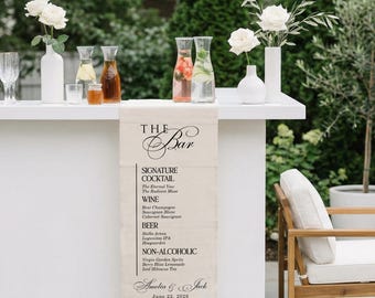 May include: A white bar with floral arrangements and various beverages. A linen bar runner displays a menu with signature cocktails, wine, beer, and non-alcoholic options. The bar is decorated for a wedding with the names Amelia & Jack and the date June 22, 2026.