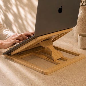 May include: A dark gray laptop with a black Apple logo on the lid is positioned on an adjustable light brown bamboo laptop stand. The stand features a slatted base and a central support arm that allows for multiple height settings for the device.
