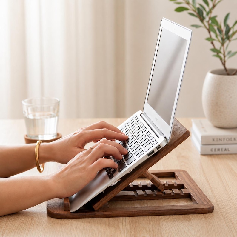 Pu&ograve; includere: Un supporto per laptop in legno scuro con un laptop argentato. Il supporto ha un design a doghe. Una persona sta digitando sul laptop. Un bicchiere d'acqua e una pianta sono sullo sfondo.