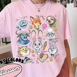 Blossom pink Comfort Colors Zootopia t-shirt with a retro character collage design. Featured characters include Assistant Bellwether, Mr. Big, and Gary De Snake in a vibrant, artistic style. This light pink oversized shirt offers a cute aesthetic for Disney World or Disneyland trips. The design is accented with pawpsicles and police badges. Ideal as a birthday gift for her, this soft cotton tee is shown in a stylish room setting with checkered shorts, making it a perfect casual summer look.
