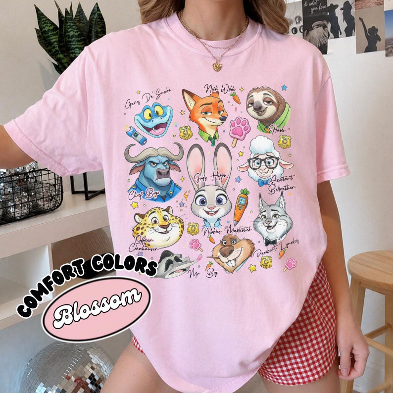 Blossom pink Comfort Colors Zootopia t-shirt with a retro character collage design. Featured characters include Assistant Bellwether, Mr. Big, and Gary De Snake in a vibrant, artistic style. This light pink oversized shirt offers a cute aesthetic for Disney World or Disneyland trips. The design is accented with pawpsicles and police badges. Ideal as a birthday gift for her, this soft cotton tee is shown in a stylish room setting with checkered shorts, making it a perfect casual summer look.