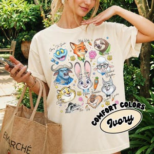 Ivory cream Zootopia character shirt in a retro 90s style. This neutral-toned Comfort Colors tee features a large collage of Judy, Nick, and the ZPD crew with playful hand-drawn accents. Perfect for a minimalist Disney park look or a cottagecore aesthetic. The soft ivory fabric pairs beautifully with the vibrant character colors. Worn by a model with a burlap tote bag in a lush garden setting. A high-quality, breathable cotton shirt that makes a thoughtful gift for movie lovers and fans.