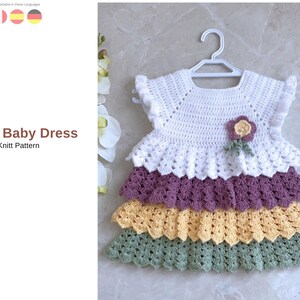 May include: A white crochet baby dress with a layered skirt in purple, yellow, and green. The dress has a floral detail and is hung on a white hanger. The text "Maggi Baby Dress" and "Knitt Pattern" are visible.