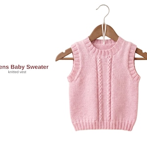 Puede incluir: Un chaleco de bebé de punto rosa claro cuelga de una percha de madera. El suéter sin mangas tiene cuello, sisas y dobladillo de canalé. El texto "Athens Baby Sweater" y "knitted vest" está a la izquierda.