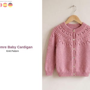 May include: A pink baby cardigan with a round neckline and button-up front, hanging on a wooden hanger. The knitted sweater features decorative patterns around the neckline and is labeled "Simre Baby Cardigan Knit Pattern".