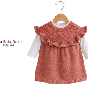 May include: A coral baby dress with a knitted vest design, hanging on a wooden hanger. The dress has a ruffled collar and a white long-sleeved shirt underneath. The text "Pera Baby Dress" and "knitted vest" are visible.