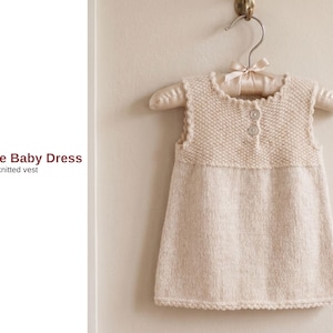 May include: A cream-colored knitted baby dress with a textured bodice and a smooth skirt, hanging on a satin hanger. The dress features two small buttons and a scalloped neckline. The text "Mepare Baby Dress" and "knitted vest" is visible.