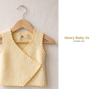 May include: A light yellow knitted baby vest with a scalloped edge, hanging on a wooden hanger. The vest has a wrap-style front and a small bow detail. Text on the right reads "Henry Baby Vest, knitted vest."
