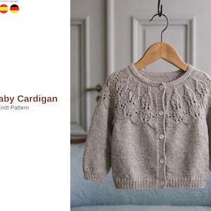 May include: A light brown knitted baby cardigan with a round neckline and button closure. The cardigan has a decorative openwork pattern across the chest and is hanging on a wooden hanger. The text "Ata Baby Cardigan Knitt Pattern" is visible.