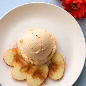 May include: A single scoop of pale beige ice cream topped with a dusting of brown cinnamon, served on a white plate with four thin, circular slices of fresh apple arranged around the base, accompanied by two vibrant red dahlia flowers.