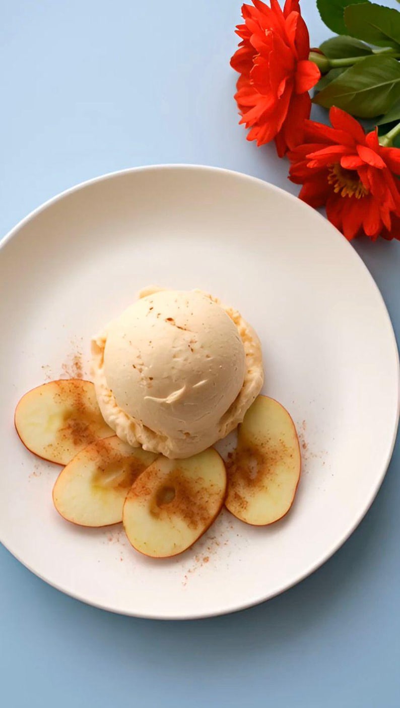 May include: A single scoop of pale beige ice cream topped with a dusting of brown cinnamon, served on a white plate with four thin, circular slices of fresh apple arranged around the base, accompanied by two vibrant red dahlia flowers.