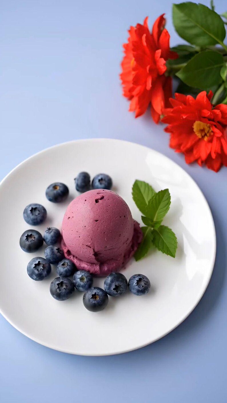May include: A single scoop of deep purple berry ice cream served on a white plate, surrounded by a cluster of fresh blueberries and a small sprig of green mint leaves, with two vibrant red flowers positioned near the edge of the plate.