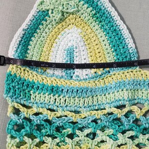 Pu&ograve; includere: Un top all'uncinetto triangolare con una sfumatura di filato bianco, giallo chiaro e verde acqua. Un metro a nastro indica una larghezza di circa 15 cm. Il design include un piccolo fiore all'uncinetto e un motivo a rete.