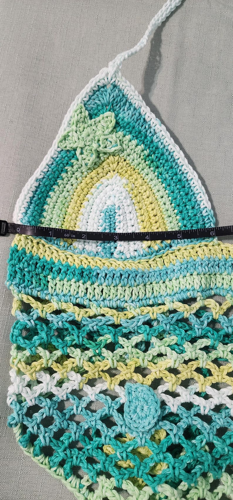 Pu&ograve; includere: Un top all'uncinetto triangolare con una sfumatura di filato bianco, giallo chiaro e verde acqua. Un metro a nastro indica una larghezza di circa 15 cm. Il design include un piccolo fiore all'uncinetto e un motivo a rete.