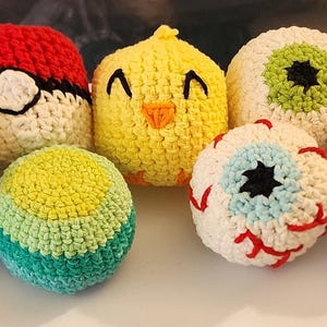 May include: A collection of seven round, crocheted yarn balls featuring various designs including a red and white ball with a black stripe, a yellow chick, two eyeball designs with red veins, a green and teal ball, a speckled beige ball, and a pastel swirl.