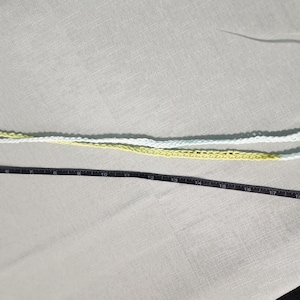 Pu&ograve; includere: Un top all'uncinetto con motivo triangolare nei toni del bianco, verde chiaro e verde acqua, con lunghe spalline all'uncinetto gialle e bianche. Un metro a nastro nero &egrave; posizionato lungo le spalline, indicando una lunghezza di circa 56 cm.