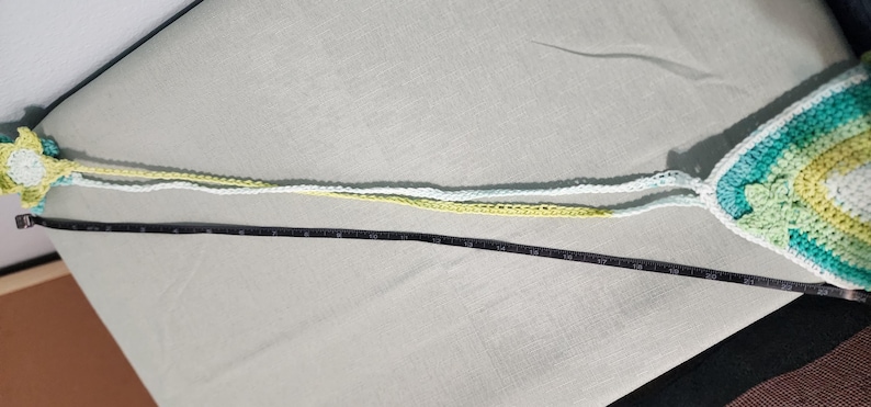Pu&ograve; includere: Un top all'uncinetto con motivo triangolare nei toni del bianco, verde chiaro e verde acqua, con lunghe spalline all'uncinetto gialle e bianche. Un metro a nastro nero &egrave; posizionato lungo le spalline, indicando una lunghezza di circa 56 cm.