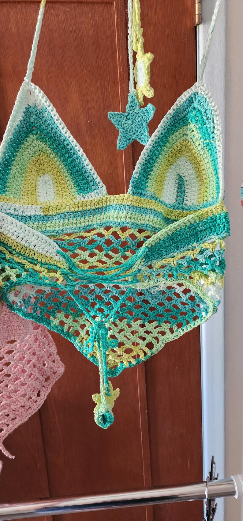 Pu&ograve; includere: Un top all'americana all'uncinetto con un design triangolare e strisce orizzontali nei toni del verde, giallo e bianco. La parte inferiore presenta un motivo a rete aperta e le spalline sono decorate con piccole stelle all'uncinetto.